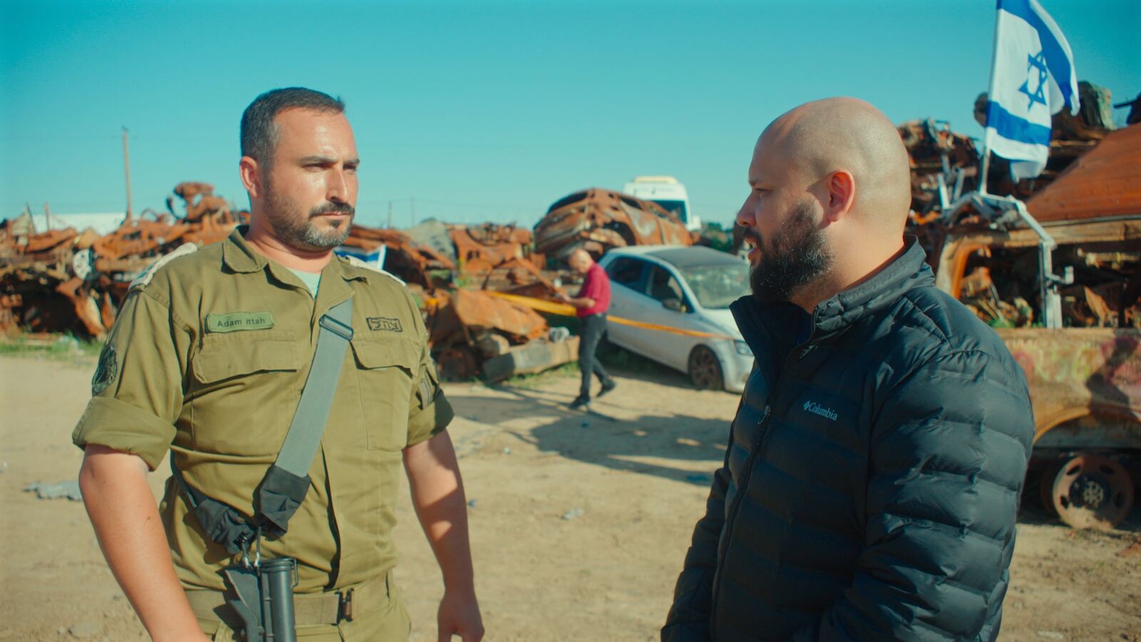 O apresentador conversa com um soldado israelense diante de carros destruídos e de uma bandeira de Israel
